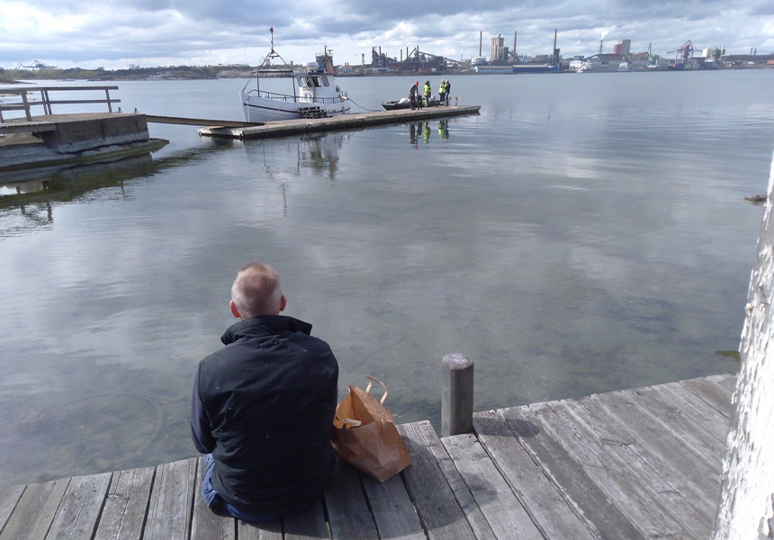 En person sitter på en brygga och tittar på SSAB Oxelösund.